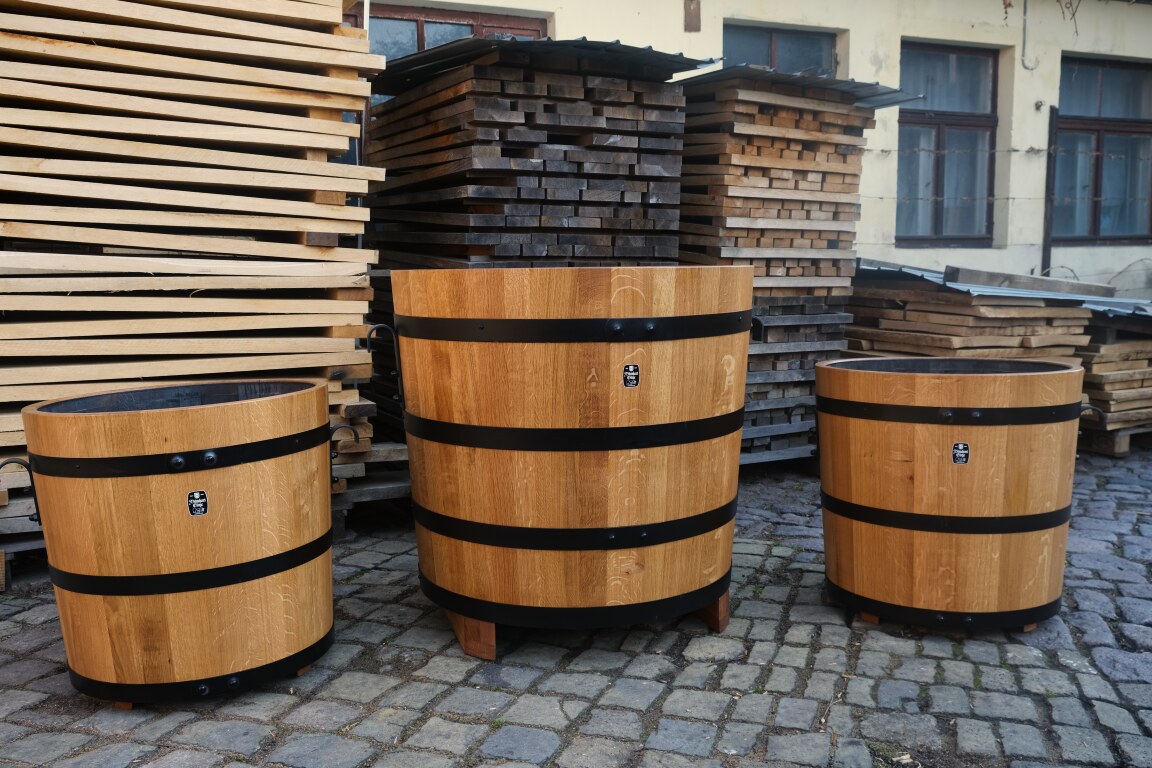 drei aufgestellte groeßere Pflanzkuebel aus Holz mit schwarz galvanisierten Reifen vor Fassholzstapeln