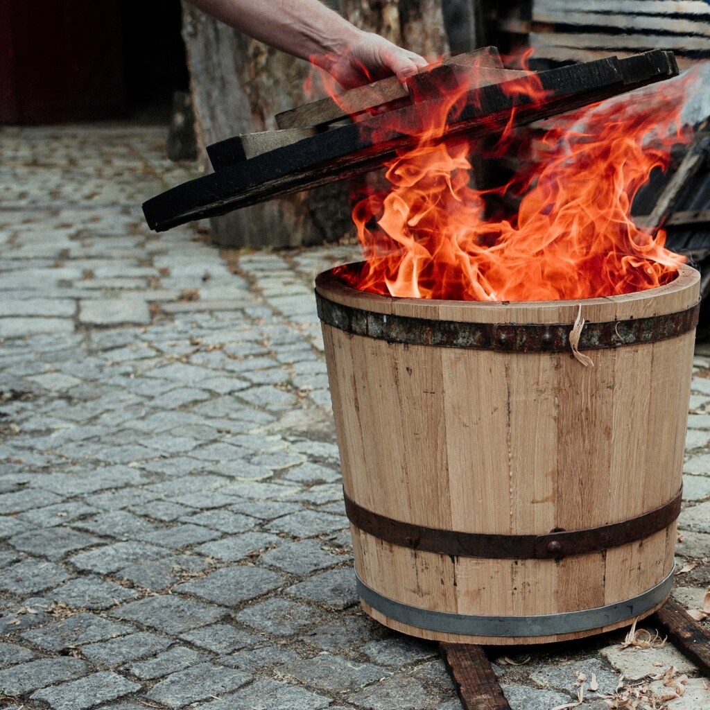 Ein Holzpflanzkuebel wird während der Produktion ausgefeuert. Er steht in Flammen. Ein Loeschdeckel ist bereits über dem Pflanzkübel.